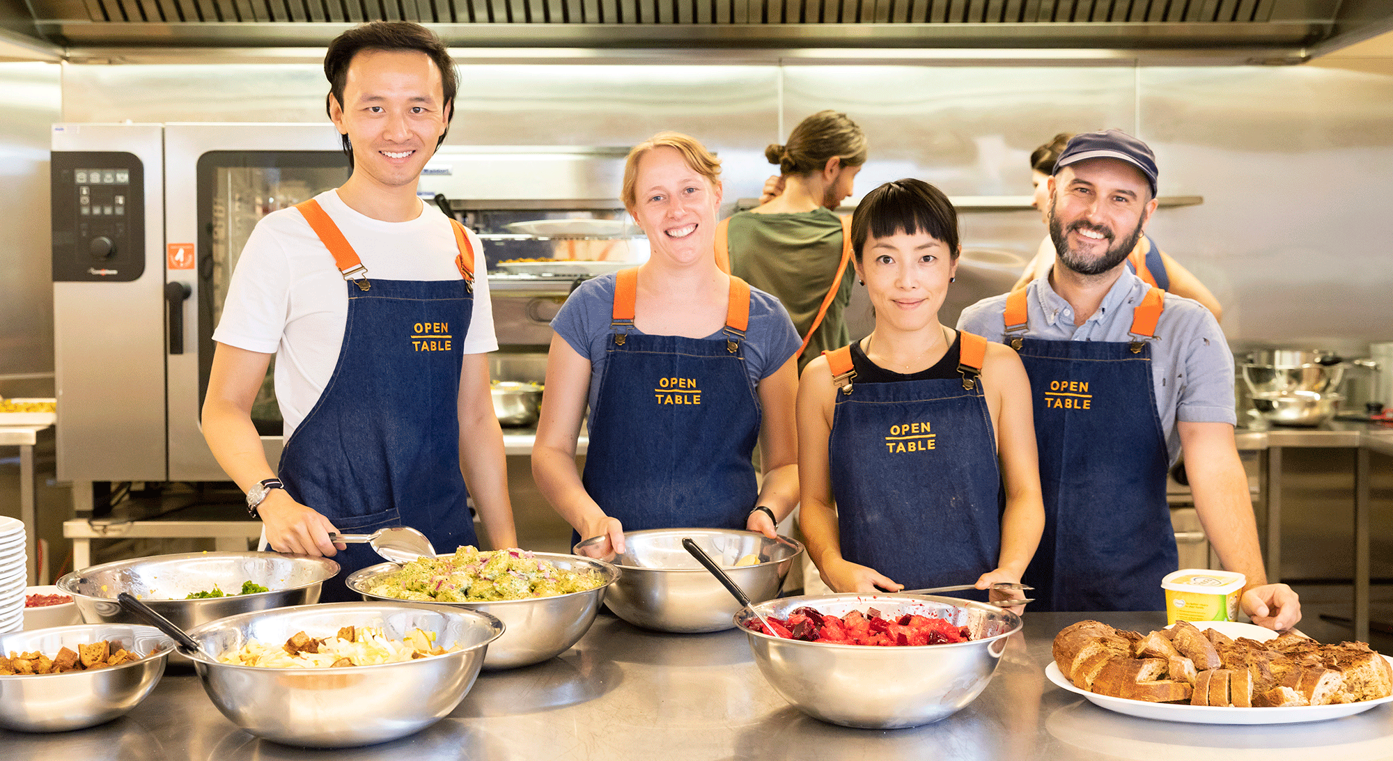 Four people wearing navy aprons that say Open Table on them standing together in a commercial kitchen