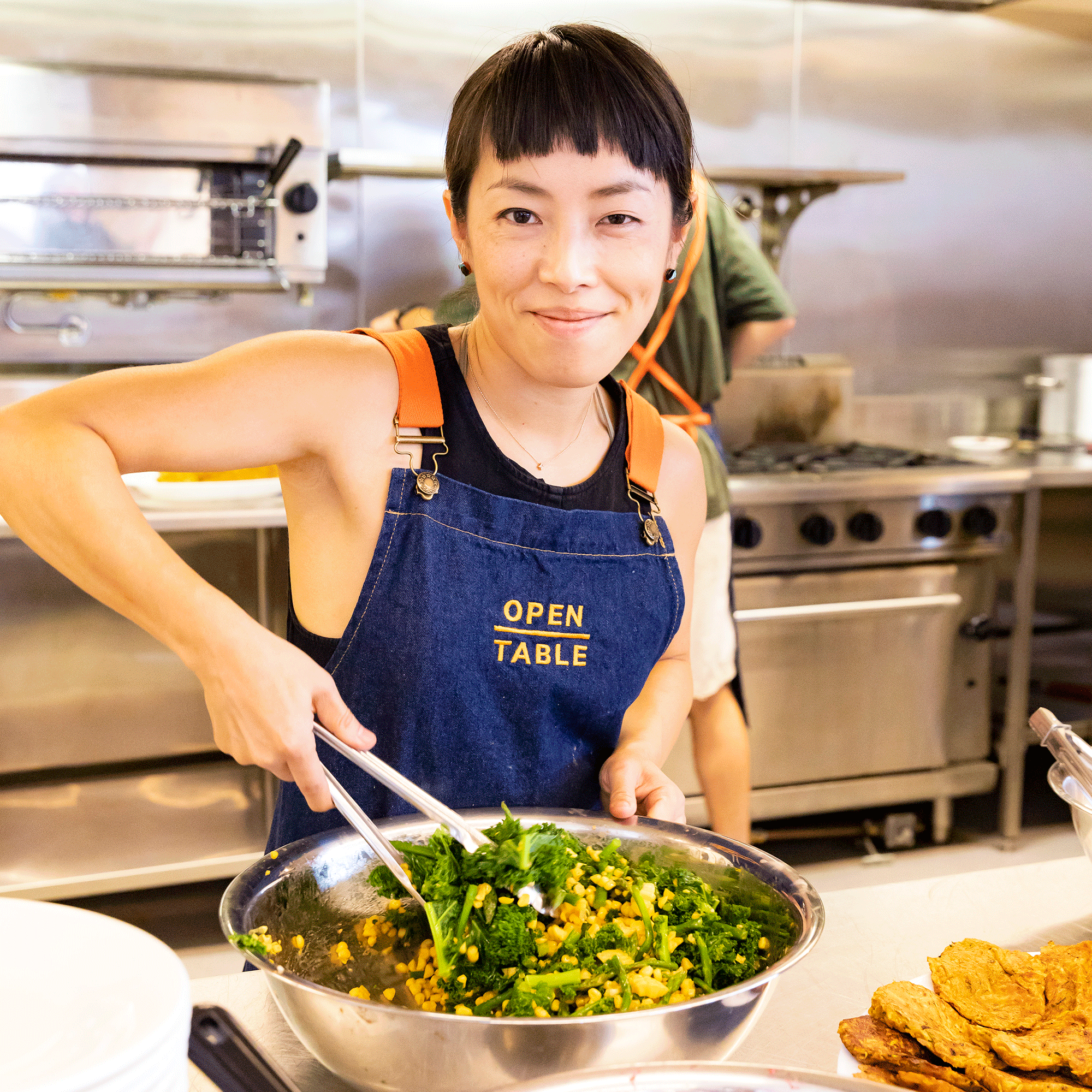 A smiling woman in a kitchen stirring a green vegetable salad 