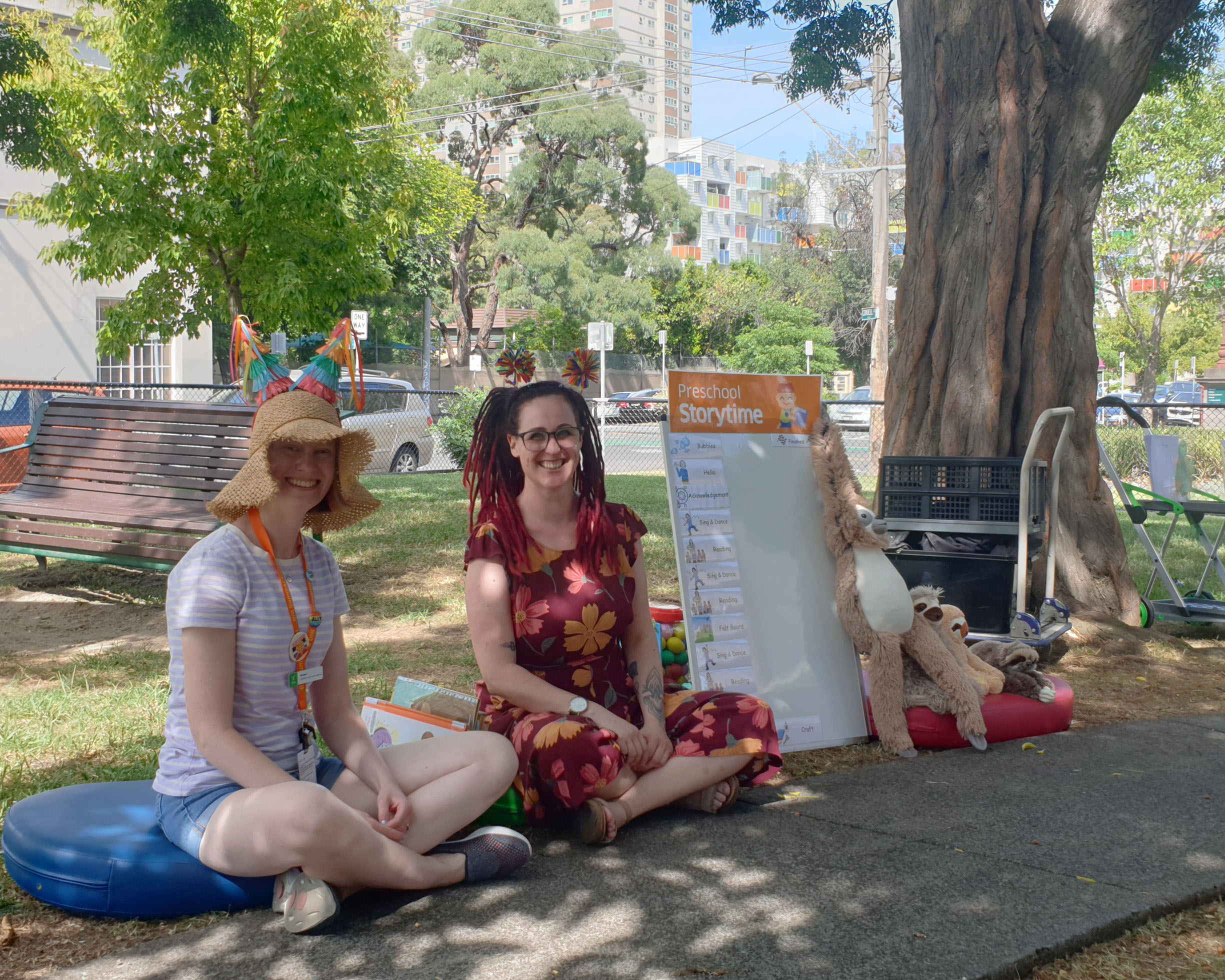 Two female librarians preparing to deliver a storytime in a sunny park with soft toys and a schedule board next to them