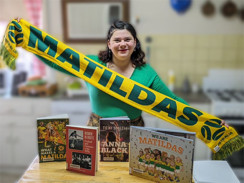 Librarian Connor holding up a Matildas scarf above a collection of women's soccer-related books