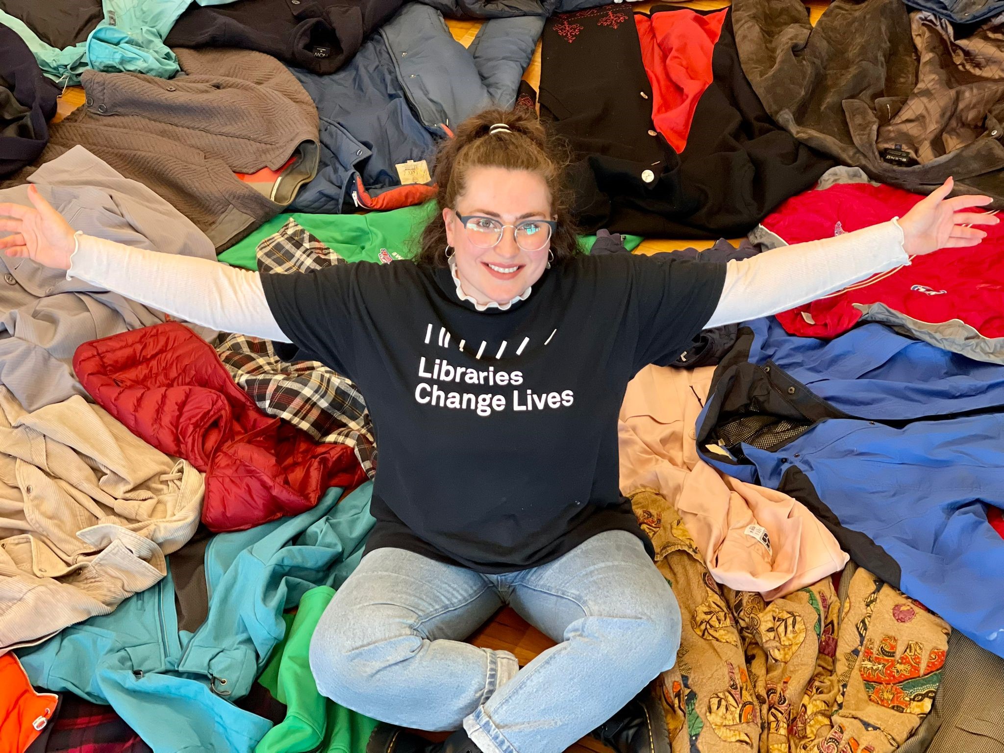 A Yarra Libraries staff member wearing a black 'Libraries Change Lives' t-shirt sits on a pile of numerous winter coats with their arms outstretched. 