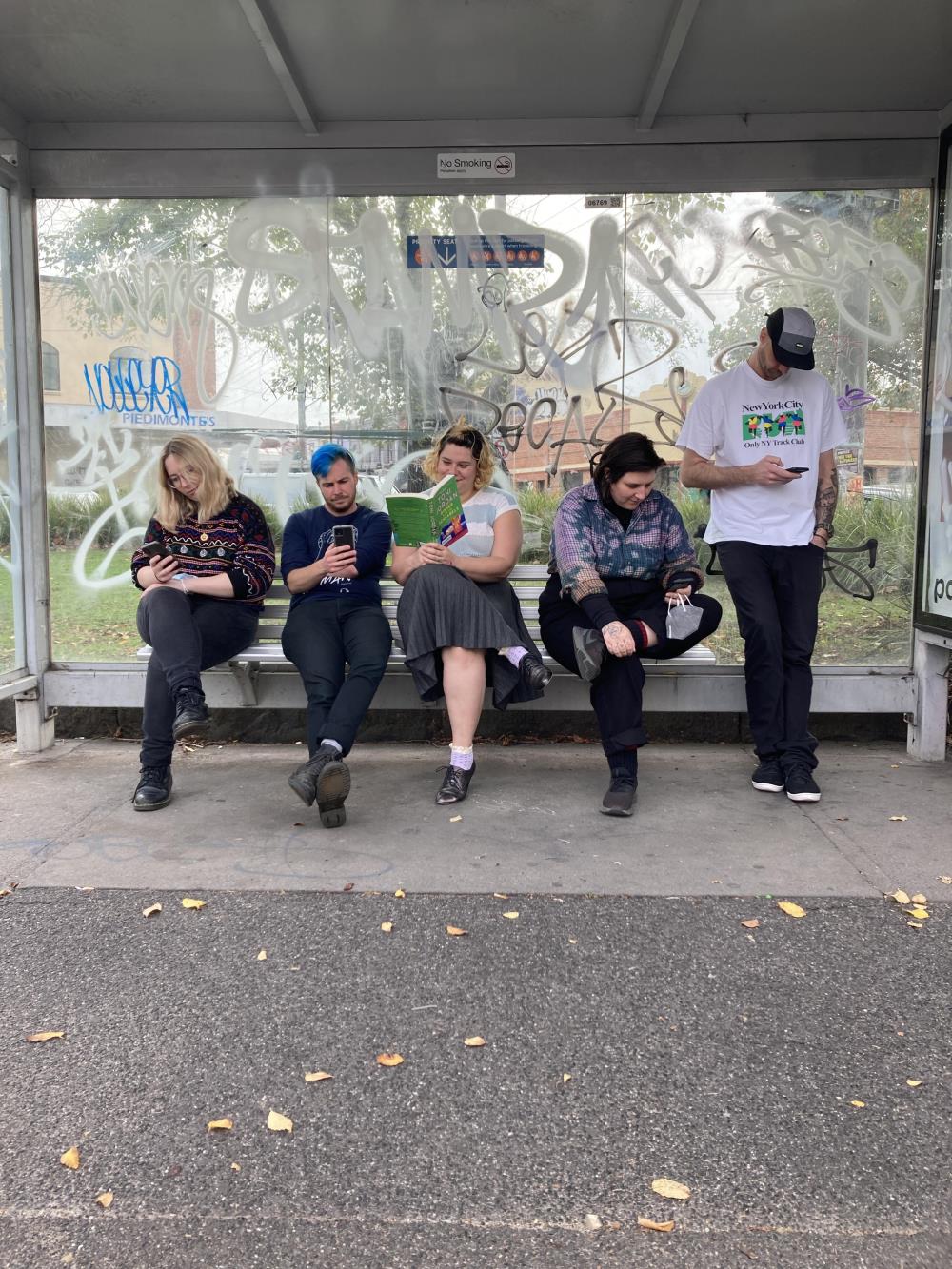 A group of five young people sit at a tram stop, all glued to their phones, except for the person in the middle who is reading a book