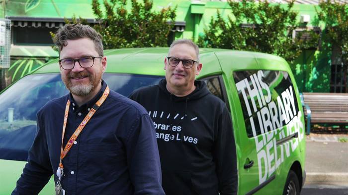Two library staff members from the Home Library Service team standing in front of the Yarra Libraries van, which is coloured bright green with the words 'this library delivers' on it. 